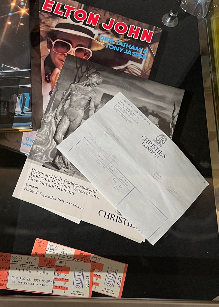 Items relating to Elton John. One is a book simply titled Elton John, with a picture of the star wearing a wide brimmed sunhat and big white glasses on the cover. That's overlaid with a Christie's catalogue entitled British and Irish Traditionalist and Modernist Paintings, Watercolours, Drawings and Sculpture. That booklet is accompanied by an invoice to F Mercury Esquire, 5-6 Logan Mews, London, W8 6QP, on which Freddie has written Gift for Elton. Finally, below those books are a couple of tickets for Sasson Presents: Elton John at the Fabulous Forum on Thursday August 23, 1984 at 8pm.