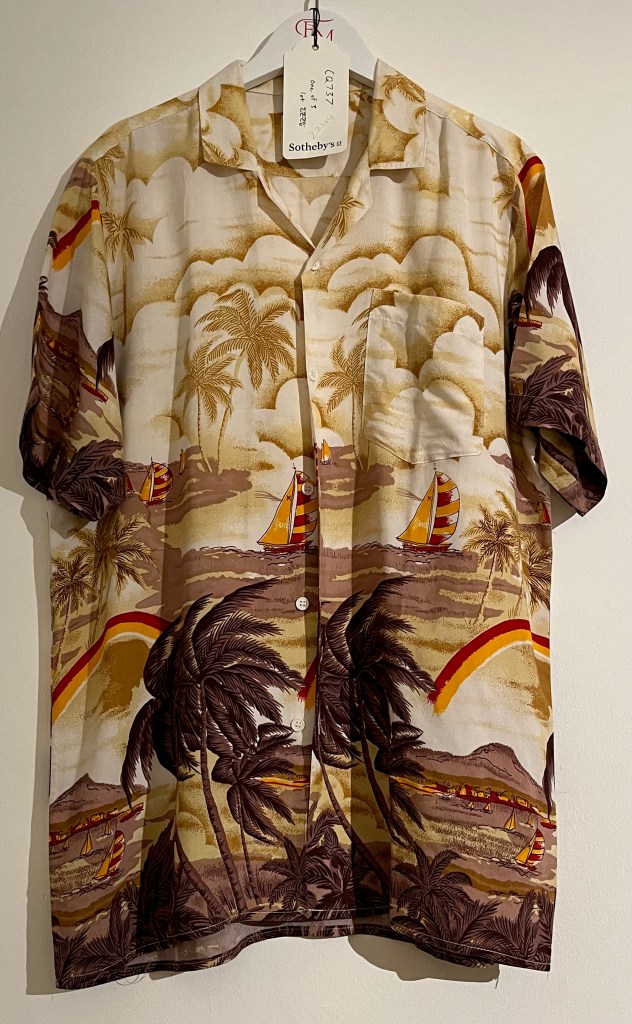A white Hawaiian shirt printed with sailing boats and palm trees beneath a cloudy sky, in yellow, red, orange and brown colours.