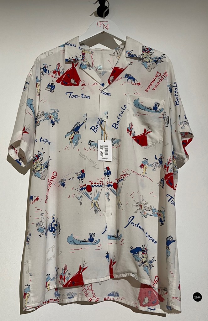 A white Hawaiian shirt printed with images of people engaging in various outdoor activities, interspersed with Native American phrases such as Indian tepee, Tom-tom, Buffalo and Chippewa drummers.
