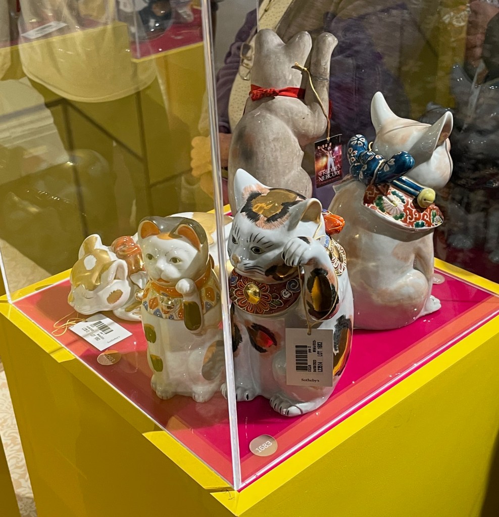 Glass case containing Japanese models of cats. At the corner nearest us are 2 cats, both seated with one paw raised, each decorated in vibrant red and blue enamels and gilt. Just behind them is a model of a cat laying down asleep, with a red ribbon around its neck.