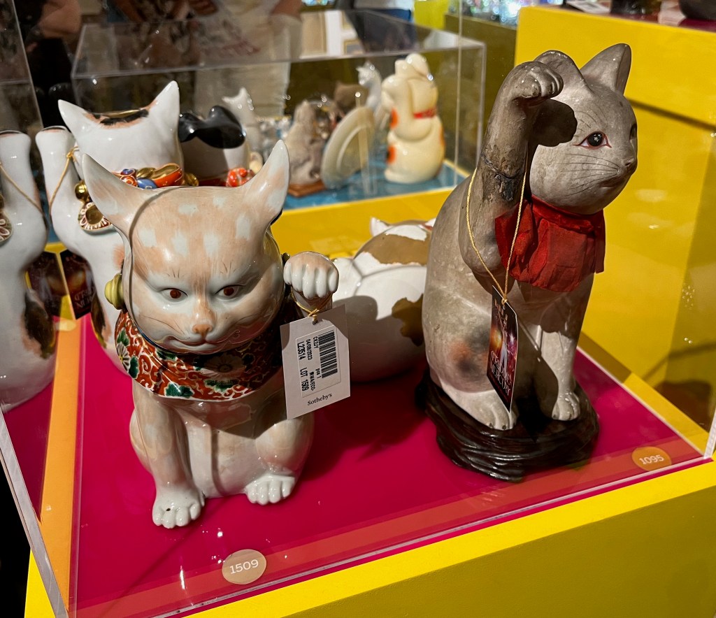 Glass case containing Japanese models of cats. The side nearest us contains two of those ornaments, each of which has a paw raised in the air. The one on the left has an elaborate brocade collar, while the one on the right is stretching up higher with its paw as if wanting the most attention of all the ornaments in the case. It has a red silk cloth around its neck and realistic detailing on its face including whiskers.