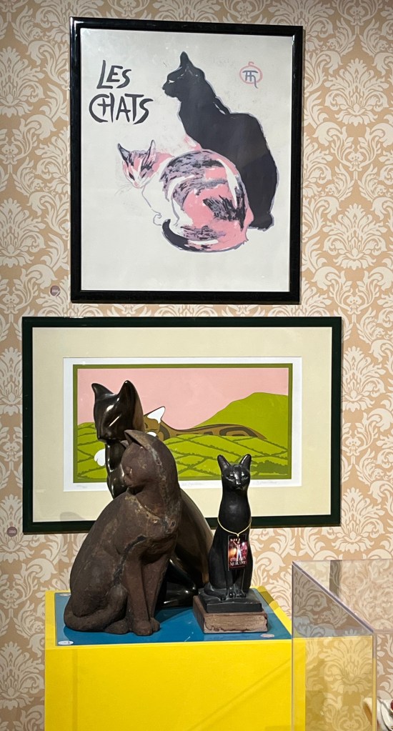 A photographic print of two cats. One is a black cat sitting up and looking off to the left, while the other has white, grey and pink colours, and is looking round towards us. At the top left of the painting, in large black capital letters on the white background, are the words Les Chats, which is French for cats. On a plinth below the print are 3 black figurines of cats.