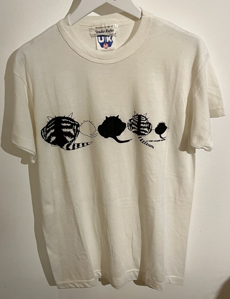 A white T-shirt printed with a row of five cats in black and white across the centre, with their backs and tails towards us.