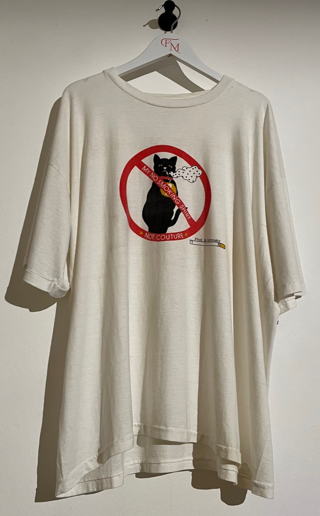 A white T-shirt printed with an image of a black cat smoking a cigarette, inside a red circle with a red diagonal line through it, like a no smoking sign. Along the diagonal line, white text says My No Smoking Shirt, while more white text on the bottom of the circle says Not Couture. To the right of the circle is a small image of a cigarette with black text above it saying Produced by Schmalor.