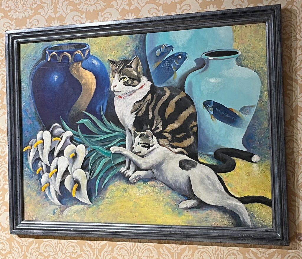 A painting of two cats. One has brown and black stripes on its back and a white chest and is sitting up attentively. The other cat is mainly white in colour except for a black spot on its back, and is laying down on the ground pawing at some long-stemmed flowers laying on the ground in front of it. Behind them are 3 large bulbous vases in varying shades of blue, a couple of which have fish designs on them.