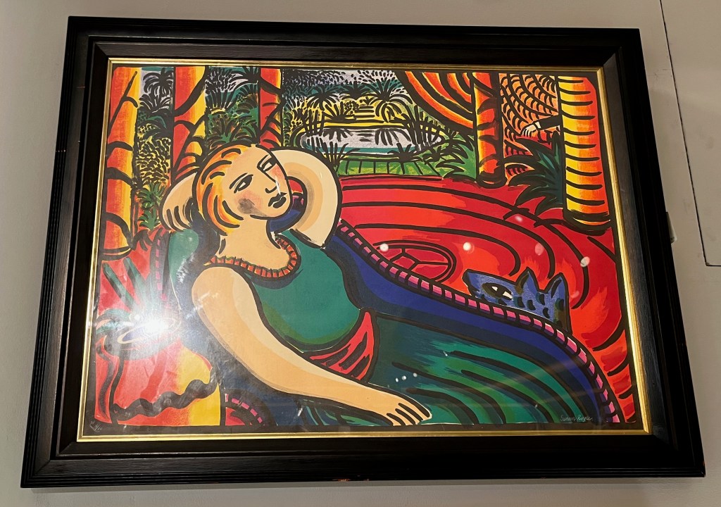 A bold, colourful painting showing a woman in a sleeveless top and a long skirt lounging on a seat, sitting back with one arm reaching around the back of her head. There are leafy plants and a pond in the background.