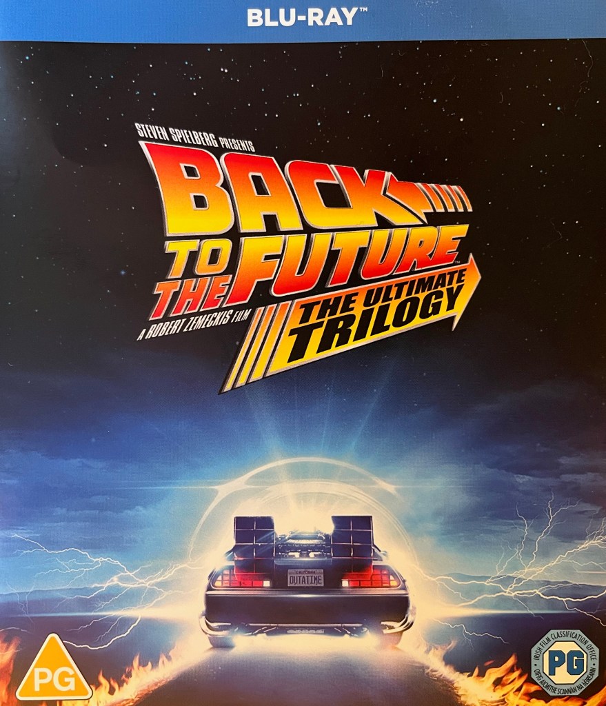 Front cover of the Blu-ray for Back To The Future - The Ultimate Trilogy. The logo for the trilogy is shown in front of a starry sky. On the ground below that, the DeLorean car, with the registration plate Outa Time, speeds towards a bright circle of light on the horizon, with flames trailing in its wake.