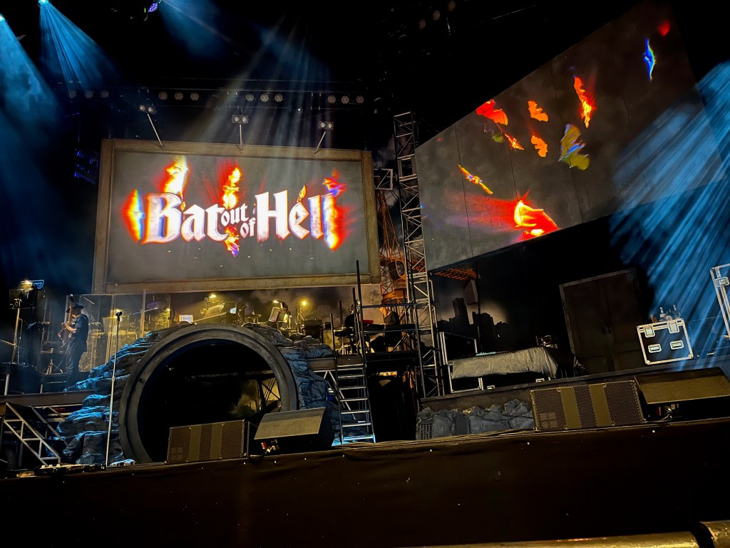 The stage for Bat Out Of Hell The Musical. On the left is a large tunnel leading off to the rear, and above the tunnel is a platform where the musicians play. On the right is a small set for Raven's bedroom. The front half of the stage is left clear. High up at the back are 2 huge screens. Both have colourful images of bats flying around, and on the left screen they're flying around the show's title, Bat Out Of Hell.