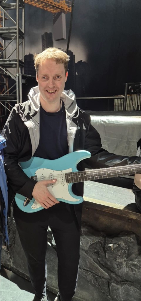 Glen smiling while holding a blue and white guitar, posing as if about to play it, with his thumb on the lowest string. He is standing on stage during the touch tour for the musical Bat Out Of Hell.
