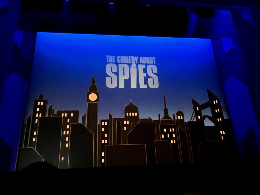The stage for The Comedy About Spies. The title of the show is in white on the back wall of the stage against a blue background, above a silhouetted and greatly simplified representation of the London skyline, including Big Ben, St Paul's Cathedral, Nelson's Column and Tower Bridge.