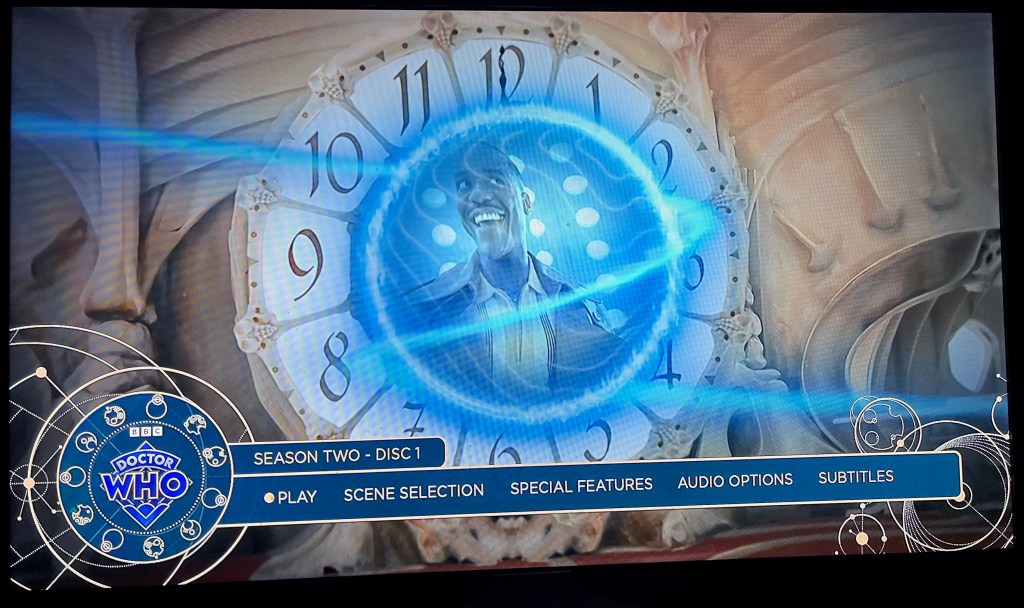 Main menu for the Blu-ray of Doctor Who Season 2, showing a large clock face, in the centre of which is an image of the Doctor smiling in the Tardis. Across the bottom are options for  Play, Scene Selection, Special Features, Audio and Subtitles.
