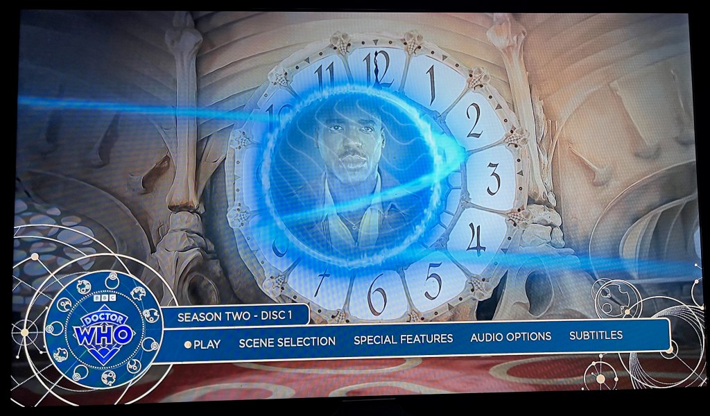 Main menu for the Blu-ray of Doctor Who Season 2, showing a large clock face, in the centre of which is an image of the Doctor looking serious. Across the bottom are options for  Play, Scene Selection, Special Features, Audio and Subtitles.