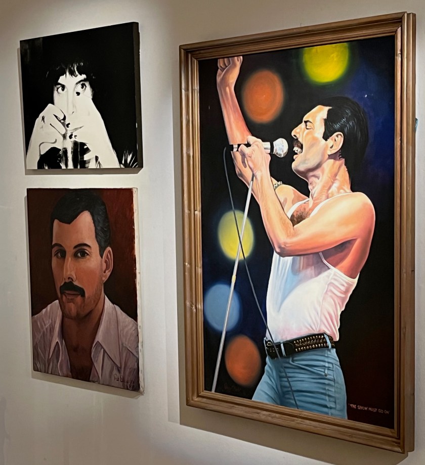 Three paintings of Freddie Mercury by his fans. Two are head and shoulder portraits, one in black and white and one in colour. The other and much larger painting shows Freddie singing loudly into a microphone as he pumps his right fist in the air, with colourful lighting behind him.