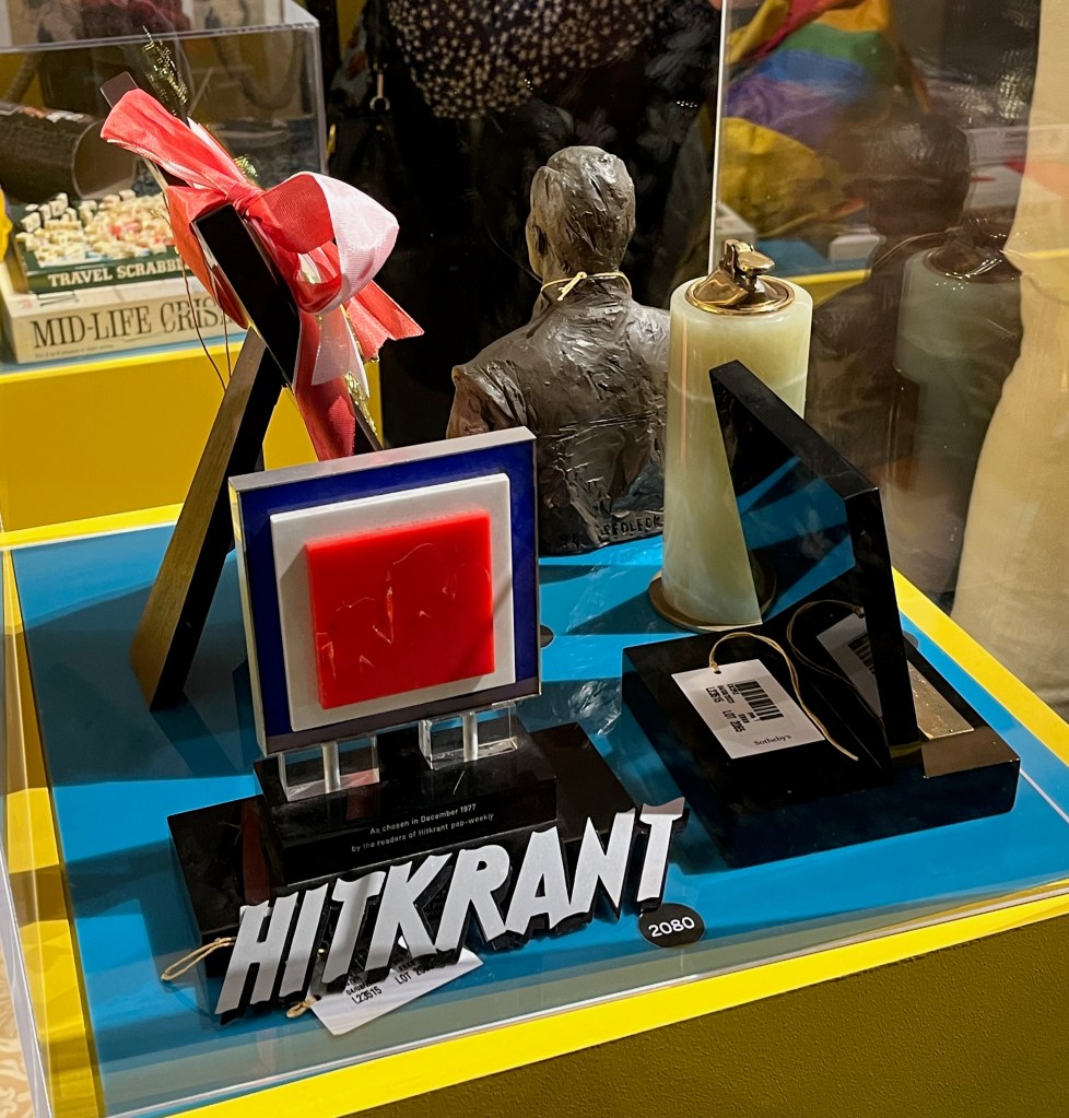Glass case with a selection of awards for Freddie Mercury. Facing us from this angle is an award consisting of concentric squares of red, white and blue from the centre outwards, and at the bottom are large letters saying Hitkrant. Elsewhere in the case is an award that looks like a very large lighter, and the back of a bust of Freddie Mercury's head and upper body.