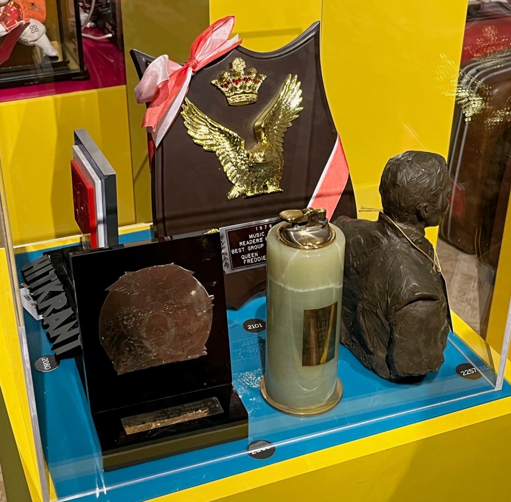 Glass case with a selection of awards for Freddie Mercury. On the left is a white metal plaque engraved with the Queen crest attached to a black resin stand. To the right of this is an award shaped like a large lighter with a brass plaque on it. Behind that is a brown resin shield with gold plastic crown and eagle motifs, decorated with red and white ribbon. And on the right of the case is a bust of Freddie's head and upper body.