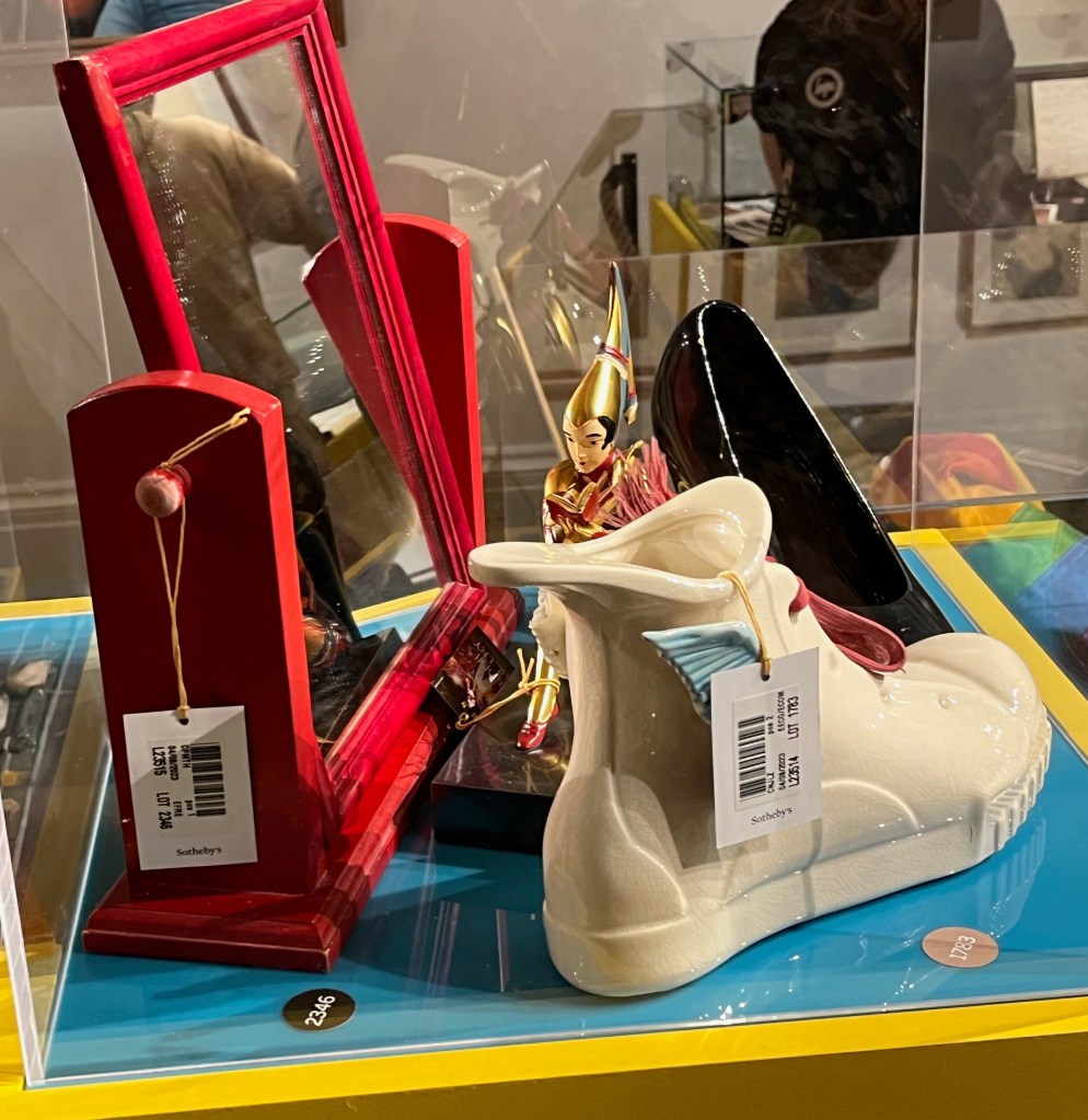 A glass case containing a red vanity mirror, a small white vase in the shape of a sneaker, a black model of a high heeled shoe, and a gold painted metal figure of a woman sitting down and reading a book.