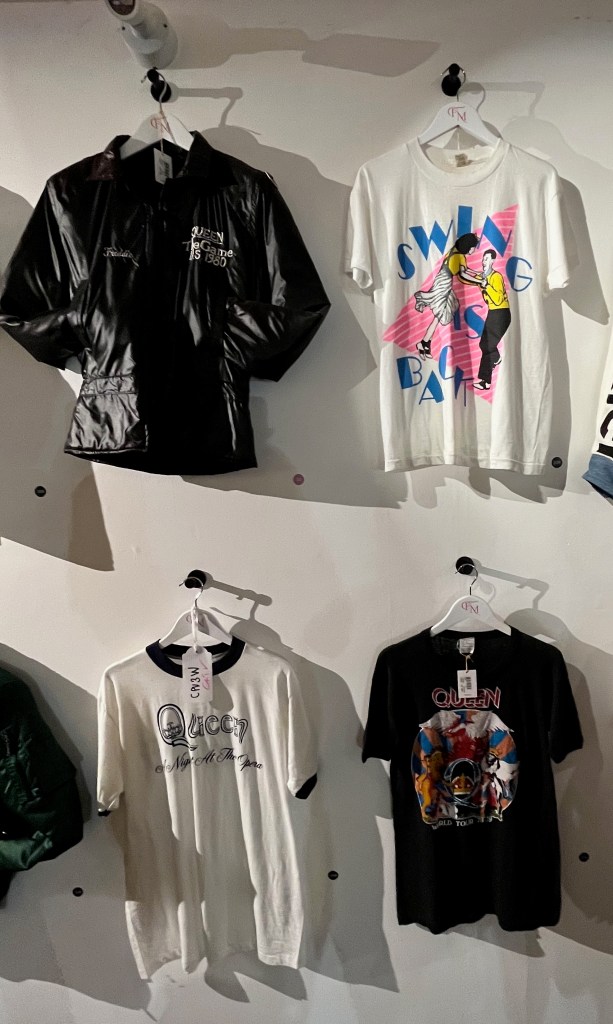 A black ‘wet-look’ nylon bomber jacket for Queen's The Game tour in the USA, a white T-shirt printed in bright colours with an image of two 1950s teenagers jiving and the text ‘Swing Is Back’, a white cotton tour t-shirt with navy trim to collar and sleeves, and navy lettering for Queen's A Night At The Opera U.S. tour 1976, and a black cotton tour t-shirt printed with red and white lettering saying ‘Queen World Tour ’78-79’ and multi-coloured designs of the Queen crest on the front.
