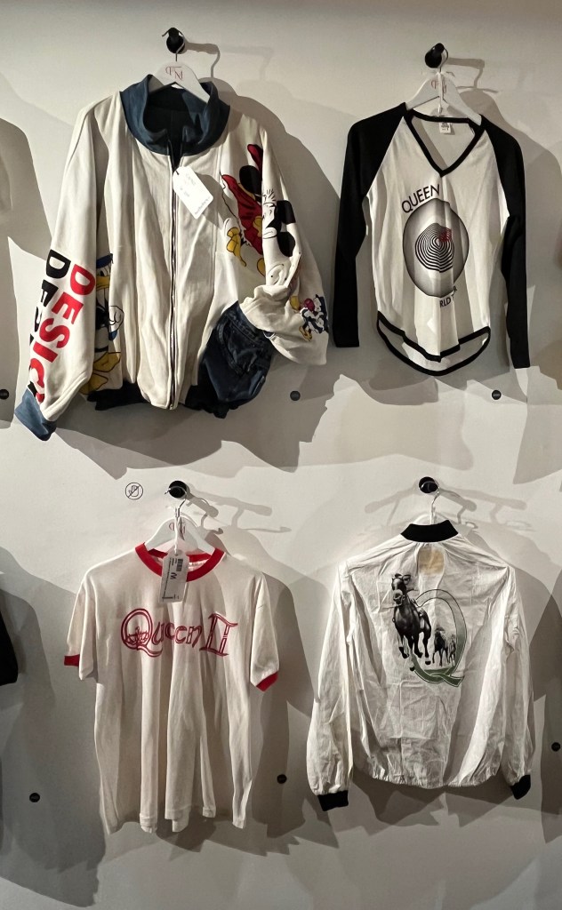 White jersey jacket printed with Disney characters Mickey and Minnie Mouse and Donald Duck, a long sleeved black and white cotton t-shirt with black sleeves and trim to v-neck and hem for Queen's Jazz world tour 1978, a white cotton tour t-shirt with red trim to neck and sleeves printed with red lettering for the Queen II U.S. Spring Tour 1974, and a white ‘Polythylene’ zip-up jacket with black knitted collar and cuffs, the reverse side shown with a black and white design of three race horses galloping through a large green letter Q.