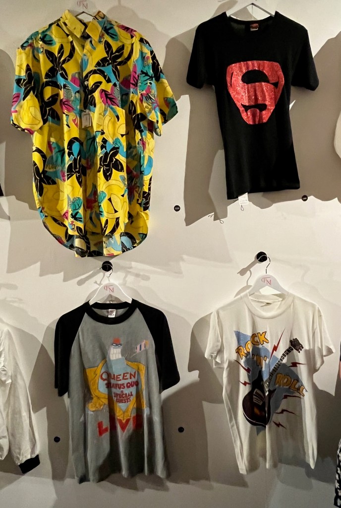A bright yellow short-sleeved Hawaiian shirt printed with an exotic floral design in fuchsia pink, turquoise and black. A black cotton t-shirt decorated with the Superman logo in red glitter. A grey cotton tour t-shirt with black sleeves and trim to collar, printed on the front with red lettering saying ‘Queen, Status Quo & Special Guests LIVE’ and a multi-coloured cartoon character from the A Kind Of Magic album cover. And a white T-shirt printed in various colours with an image of a guitar and text saying ‘Rock N Roll’.