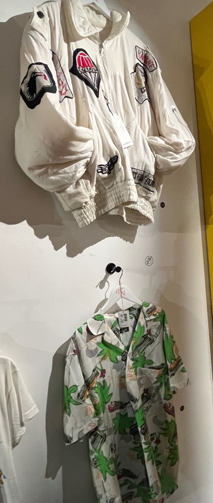 A white padded cotton jacket applied to front, back and sleeves with cotton badges embroidered in black, red and white with various Japanese characters, symbols or themes. And a white short-sleeved Hawaiian shirt printed with a design of bright green palm trees and orange and pink vintage cars.