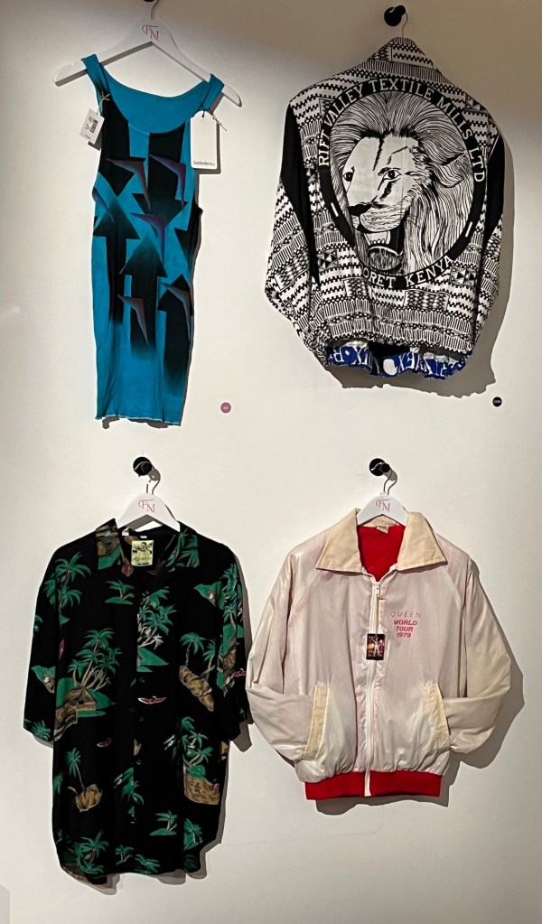 Turquoise vest with large arrow designs, with 3 pointing up and 3 pointing down, meeting in the centre, in black, purple and red, for Queen's 'Hot Space' tour 1982. A batwing sleeve cotton jacket with shoulder pads, printed with a black and white geometrical pattern, the back shown with a large lion’s head print in the centre. A black short-sleeved Hawaiian shirt printed with a pattern of palm trees, huts, sails and canoes in green, red and brown. And a white nylon bomber jacket with red knitted cuffs and hem and red quilted lining, with pink printed lettering saying ‘Queen World Tour 1979’.