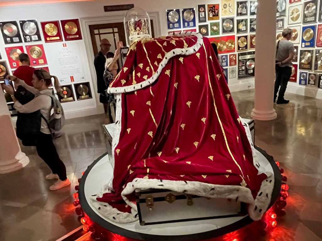 A huge royal red cloak with gold decorations and white fur trim, accompanied by an ornate gold crown, on display in the Freddie Mercury exhibition, with people keenly taking photos of it from mall angles.