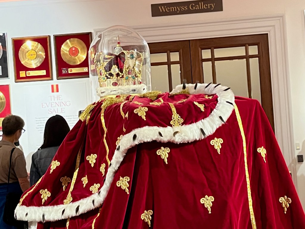 Close-up of the top section of a royal red cloak with gold decorations and white fur trim, accompanied by an ornate gold gem-studded crown with white fur trim, on display in the Freddie Mercury exhibition.