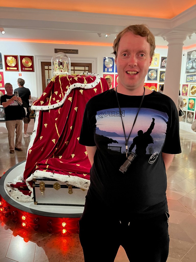Glen smiling as he stands in front of Freddie Mercury's cloak and crown ensemble. It consists of a huge royal red cloak with gold decorations and white fur trim, accompanied by an ornate gold crown. Glen is wearing a black t-shirt on which is the cover of Queen's Made In Heaven album, which shows Freddie Mercury's statue at Montreux in Switzerland, looking out over Lake Geneva.