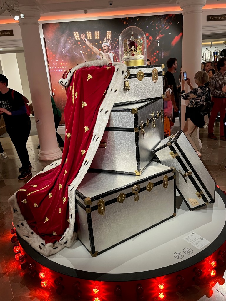 A set of large aluminium travelling trunks and suitcases, with gilt-brass locks and decorations, and black plastic riveted edging, in the Freddie Mercury exhibition. Draped over them is a huge royal red cloak with gold decorations and white fur trim, accompanied by an ornate gold crown.