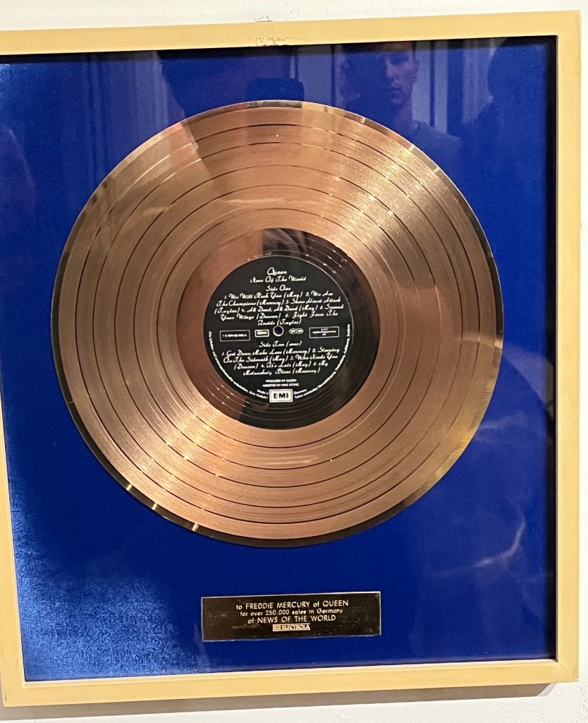 A large framed gold disc against a blue background, with a silver plaque beneath that says To Freddie Mercury of Queen for over 250,000 copies of News of the World. In the centre of the disc is a black label with white curly text listing the tracks on the album.