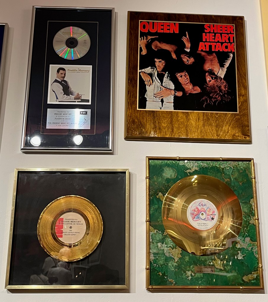 Disc awards for The Freddie Mercury Album and the Queen songs Crazy Little Thing Called Love and We Are The Champions, along with a large copy of the Sheer Heart Attack album artwork, on wood lacquer. The artwork shows the 4 members of the band lying on their back in different directions next to each other.