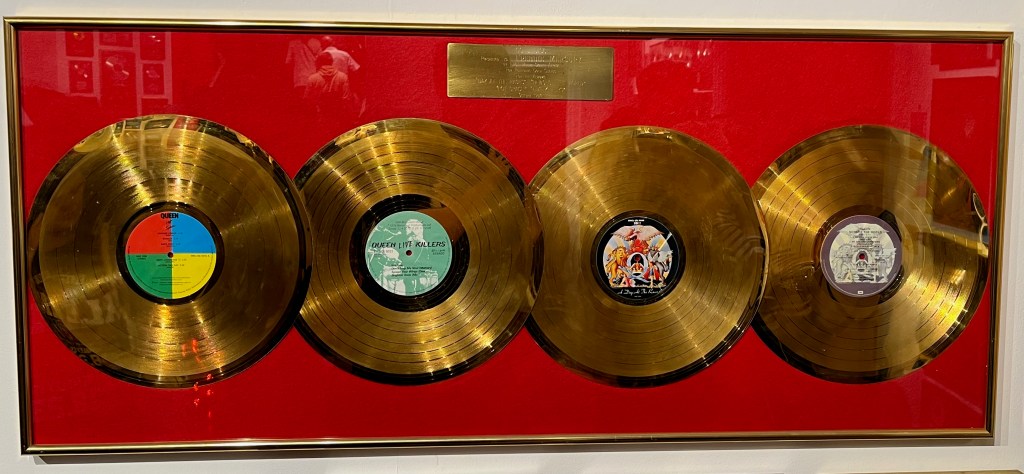 An award with 4 gold discs against a red background for the Queen Albums Hot Space, Live Killers, A Day At The Races and News Of The World.
