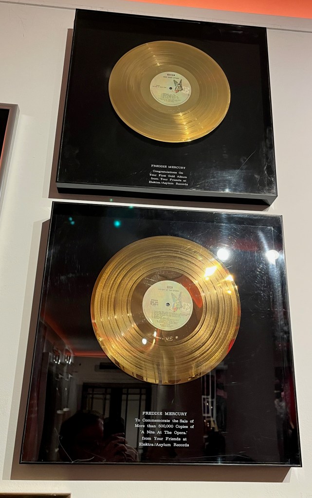 2 large gold discs on black backgrounds in perspex boxes. Each has a black plaque with white text at the bottom. The top one, for the Sheer Heart Attack album, says Freddie Mercury, congratulations on your first gold album, from your friends at Elektra Asylum records. The plaque for the bottom disc says Freddie Mercury, to commemorate the sale of more than 500,000 copies of A Night At The Opera, from Your Friends at Elektra Asylum Records. The word Night is misspelt as N-I-T-E.