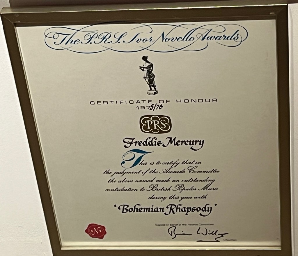 An Ivor Novello Certificate of Honour in an elaborate gilt frame, presented to Freddie Mercury for the song Bohemian Rhapsody. The text, mainly in a curly script font, reads The P.R.S. Ivor Novello Awards Certificate of Honour 1975/76, Freddie Mercury. This is to certify that in the judgment of the Awards Committee the above named made an outstanding contribution to British Popular Music during this year with Bohemian Rhapsody.