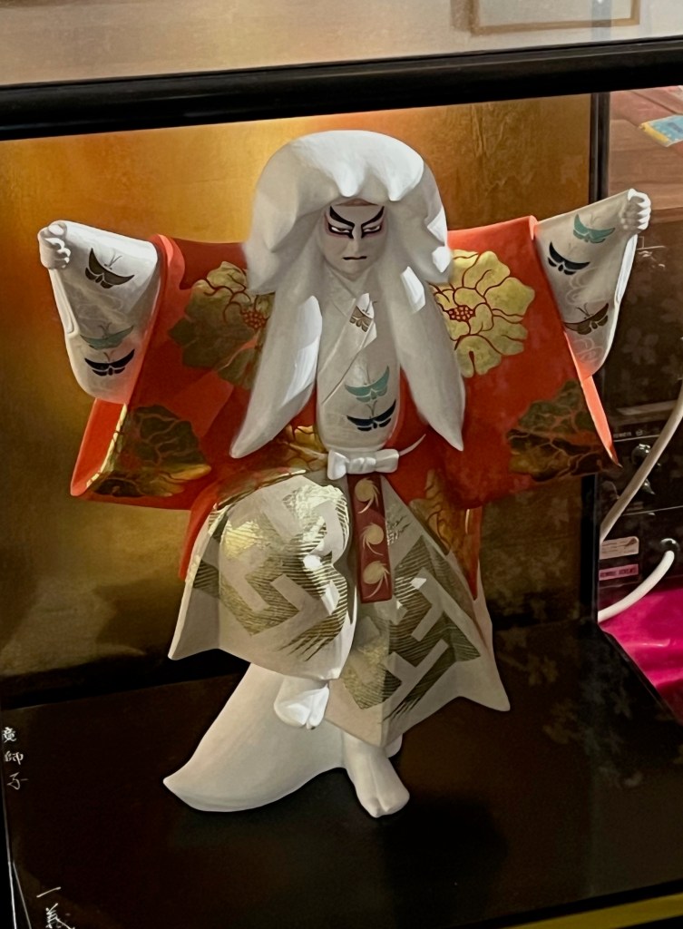 A small white figurine of a Japanese actor, wearing a colourful red, white and gold outfit decorated with peonies and stylised butterflies. His arms are outstretched to the sides, and the heavy black lines emphasising his eyes and eyebrows gives him a fierce-looking expression.