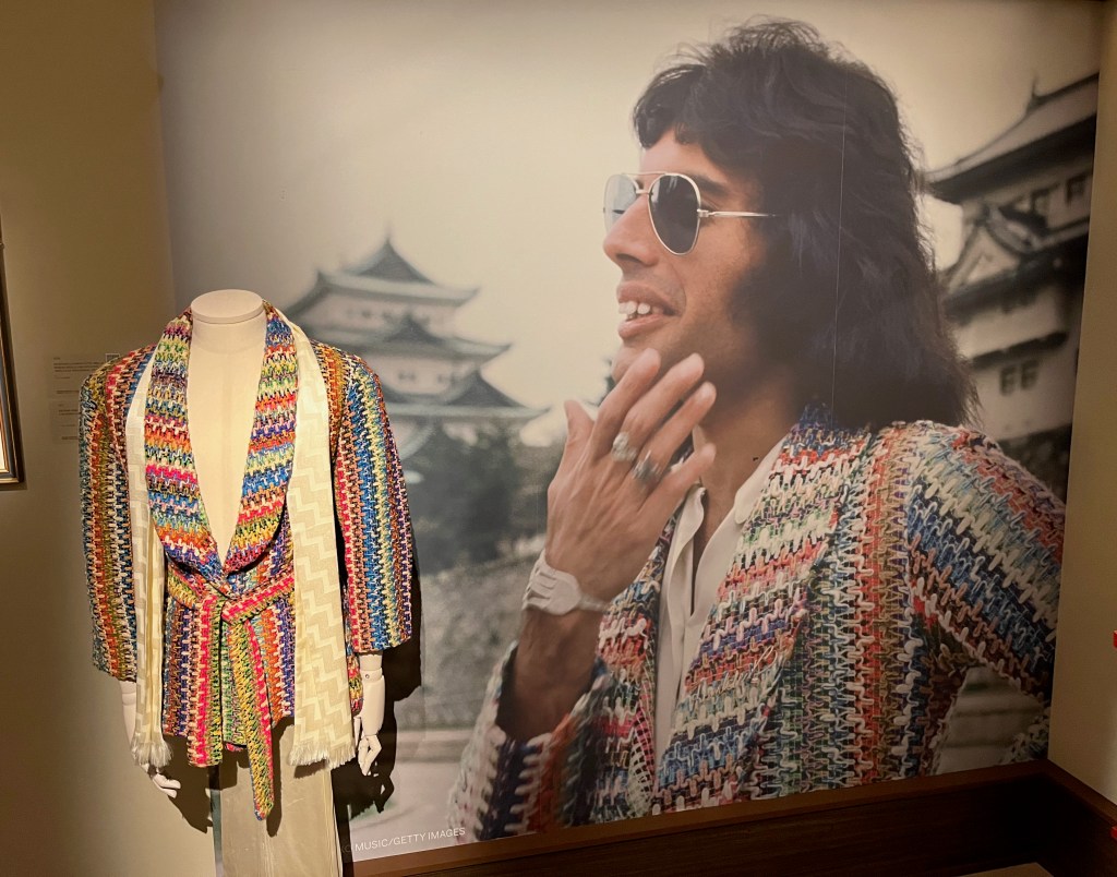 A loose-fitting brightly multi-coloured jacket with a weaved pattern, patch pockets at the hips and a matching self-tie belt. Behind the jacket is a large photo of Freddie Mercury wearing it in Japan.