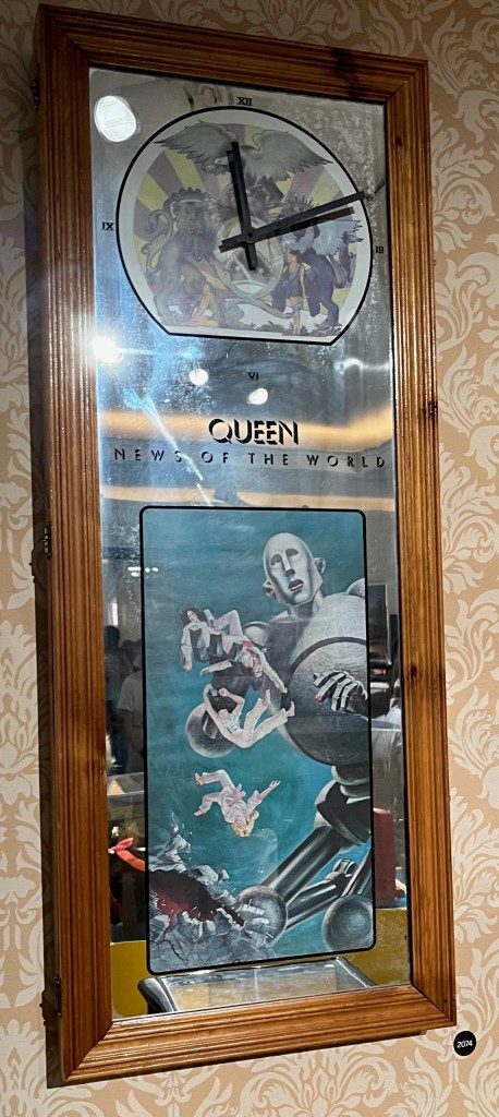 A tall mirrored walk clock. At the top is the crest for the band Queen, over which are the clock hands, and there are Roman numerals for each quarter of the hour. The centre of the item has the words Queen, News Of The World. Then the bottom half shows the artwork from the album, with a huge robotic figure picking up and dropping the band members, who are tiny in comparison.