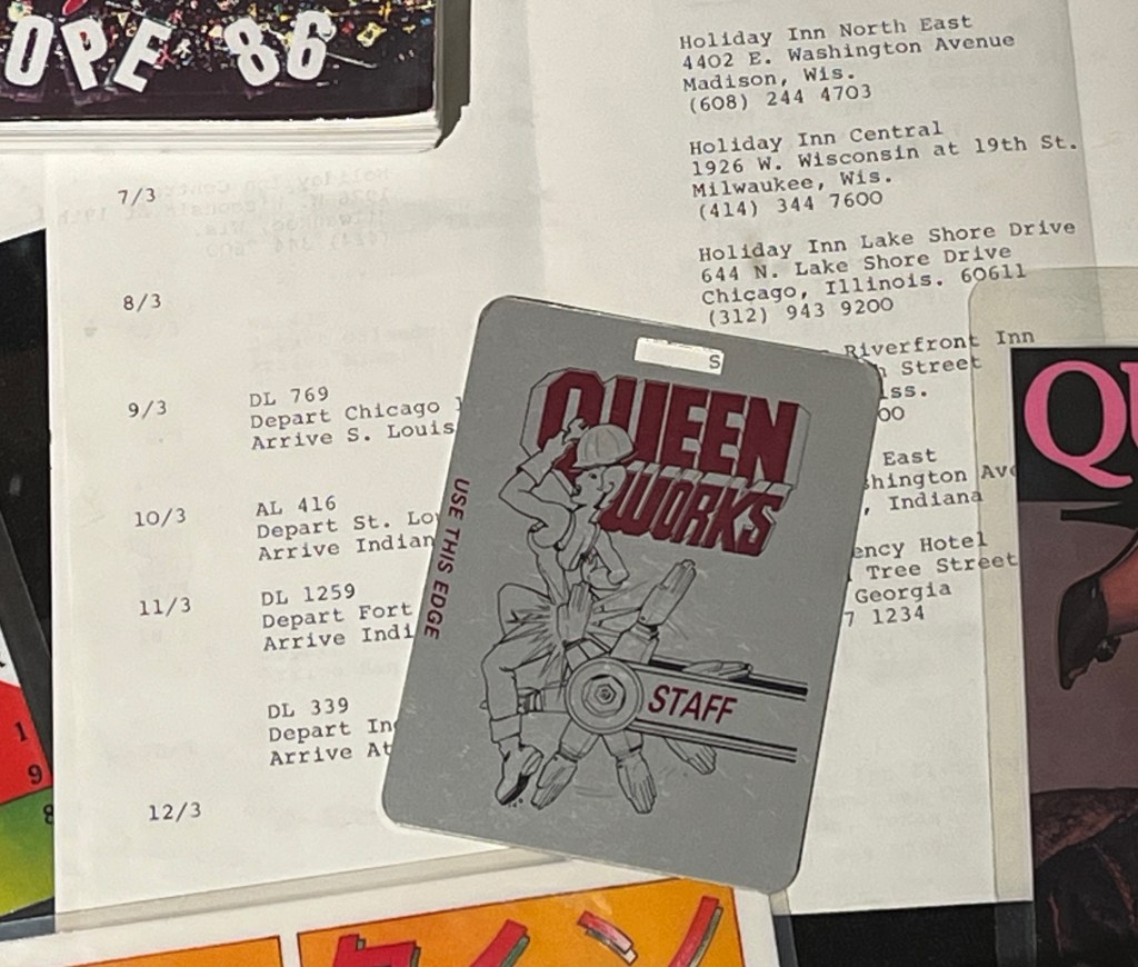 A metal staff pass for the Works tour by the band Queen. The artwork on it shows a woman wearing a hard hat and working overalls while holding a spanner, and is being slapped on the behind by a spinning wheel of hands.