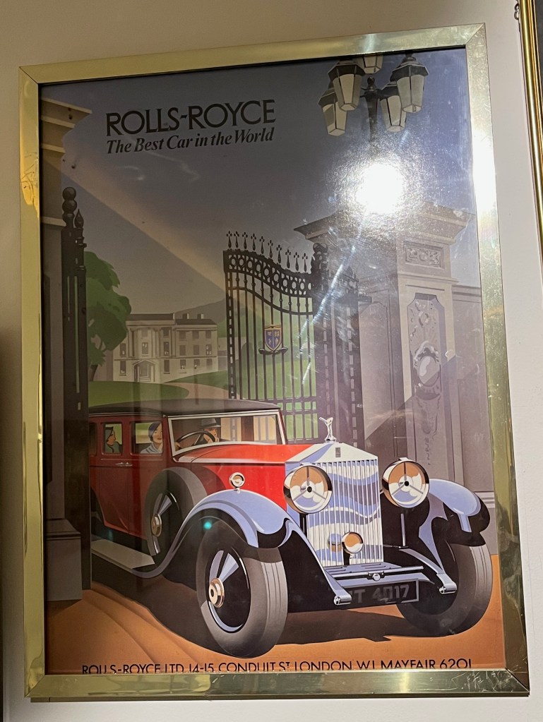 A poster showing a family driving a large red Rolls Royce car out of a big set of metal gates. Behind them is a curving path, lined with grass on either side, leading to a mansion. Text at the top of the poster says Rolls Royce, the best car in the world.