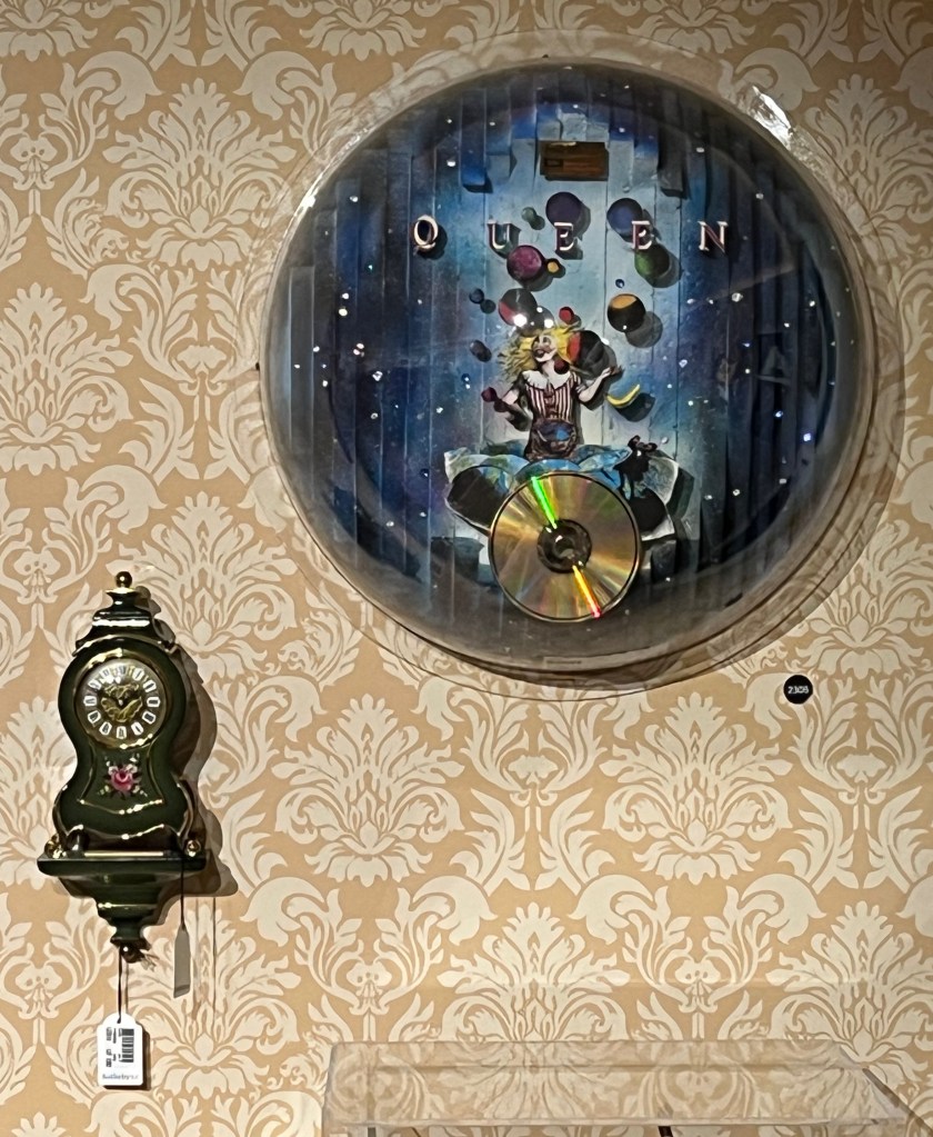 A large round award presented to Queen and Freddie Mercury for the Innuendo album. Against a dark blue background it shows the album artwork, with a smiling figure juggling, in 3D, decorated with imitation gems and silver stars, above a copy of the CD. Below this and to the left on the wall is a much smaller cartel style clock, painted with roses on an olive green background.