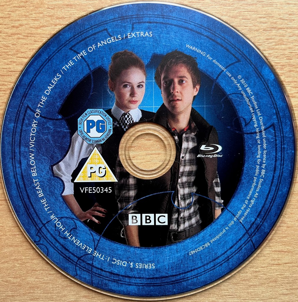 Disc 1 in the Doctor Who Series 5 Blu-ray set. It features an image of Amy Pond dressed in her police uniform, alongside Rory Williams in a chequered shirt and dark jacket. A blue border around the edge of the disc contains the episode names and copyright information.