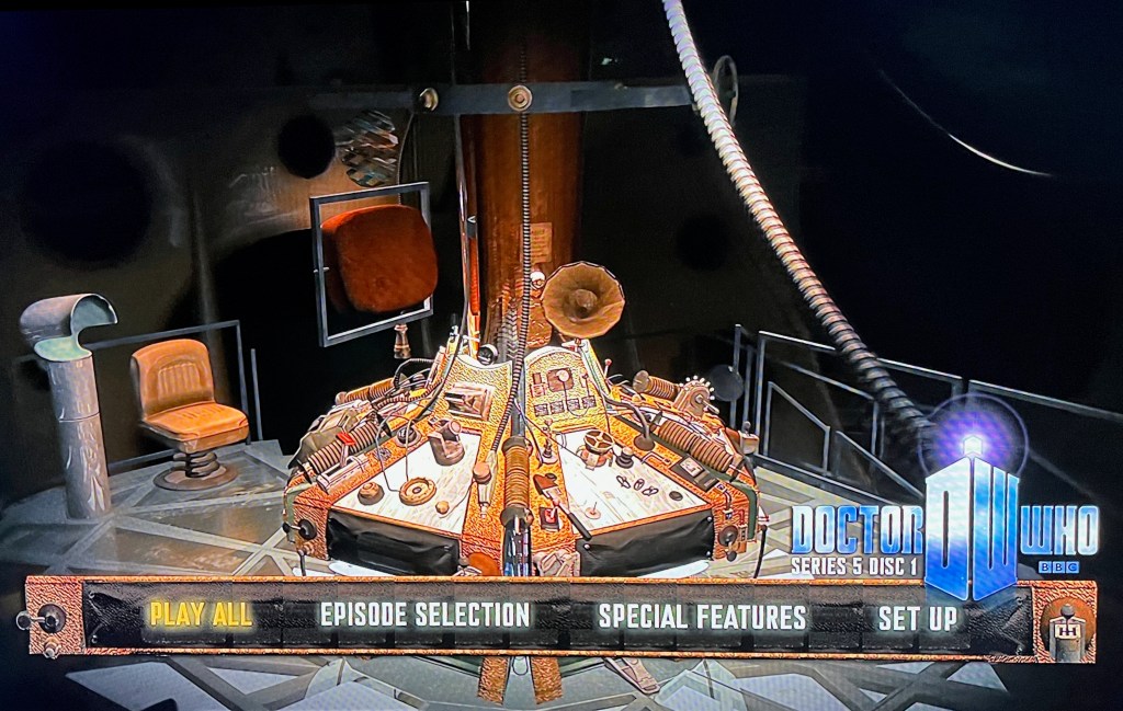 Blu-ray menu for Doctor Who Series 5, showing a full view of the Tardis console, which is covered with all sorts of buttons, dials and gadgets, and even a large round speaker horn from an old gramophone. The back of the Tardis monitor is also visible, with the front of it facing towards a chair to the left of the console. There are options along the bottom for Play All, Episode Selection, Special Features and Set Up, followed by the Doctor Who logo.