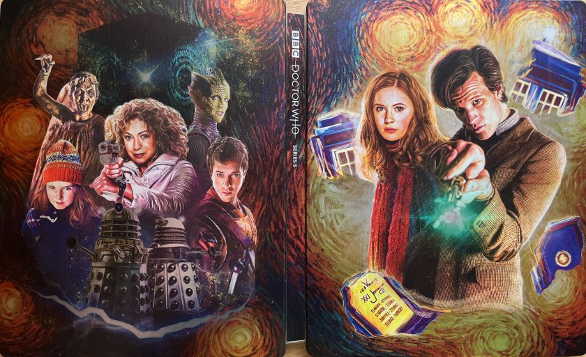 The cover spread artwork for the Doctor Who Series 5 Blu-ray steelbook. The front cover, on the right, shows Matt Smith as the Doctor, with a stern expression on his face as he points his sonic screwdriver straight at us. His companion Amy Pond, with red hair and a red scarf over her dark clothing, stands next to him, also looking towards us. In the background, the Tardis has split in two amongst a swirling explosion. The back cover, on the left, shows various other characters from the series. River Song is the largest in the centre, pointing a gun ahead of her. Surrounding her are Amy Pond as a child, a Weeping Angel, the Pandorica box, Silurian Madame Vastra, companion Rory Williams, and two Daleks.
