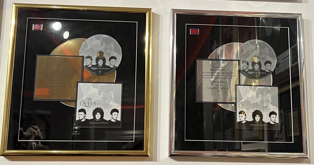 Two sales awards for Queen's Greatest Hits 3. Each award is a large box with a black background. A large gold or platinum disc sits in the centre of the background. Overlaying this on the top right is the CD, and below that the album cover, both carrying an image of Freddie singing and looking down over the other 3 band members who are in a line. In a central position to the left of the disc and album art is a gold or platinum plaque explaining why the award has been given. The plaques say the awards are presented to Mary Austin in honour of Freddie Mercury.