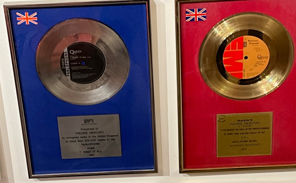Large disc awards in display boxes for Queen singles, including a silver disc on a blue background  for I Want It All, and a gold disc on a red background for Bohemian Rhapsody