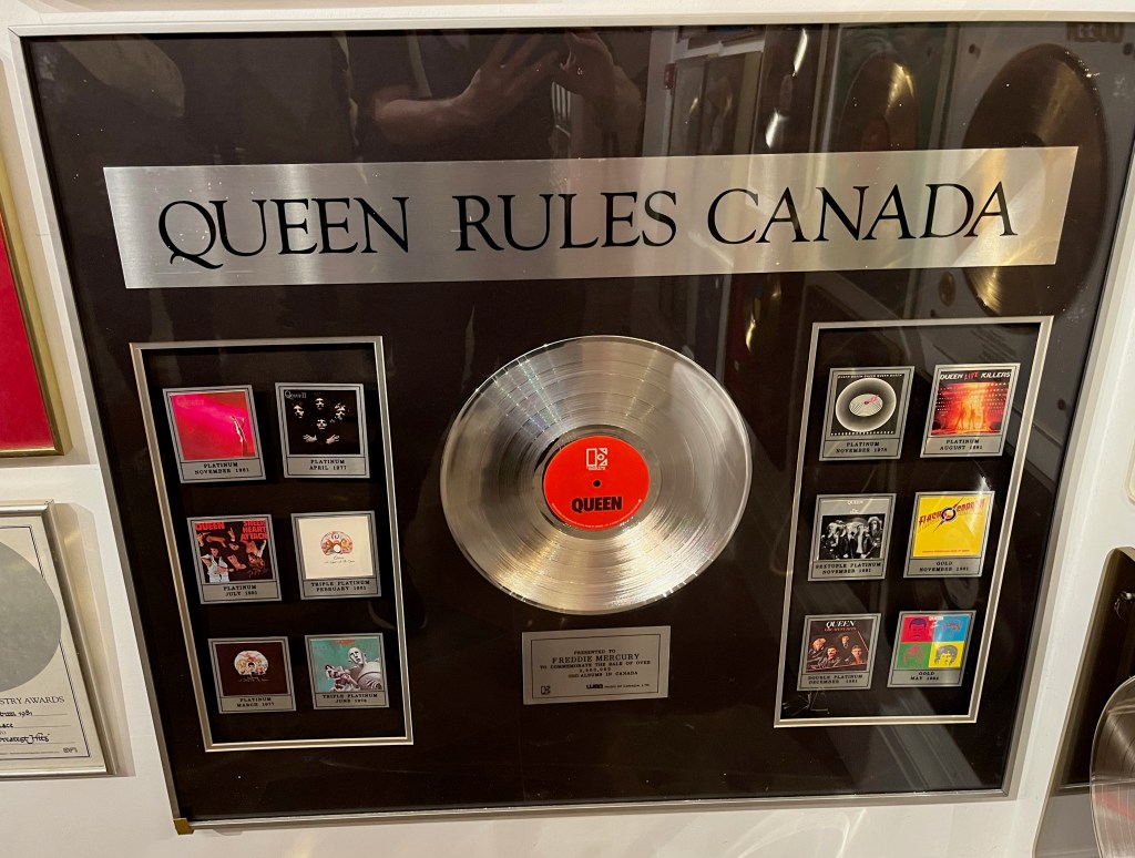 A large display case with a black background. At the top, a large silver strip with black letters says Queen Rules Canada. Below it, in the centre, is a silver disc with a red centre that has the word Queen in black letters. Either side of the disc are images of Queen's album covers, 6 per side, from Queen to Hot Space. A silver plaque below the central disc says Presented to Freddie Mercury to commemorate the sale of over 2,500,000 Queen albums in Canada.