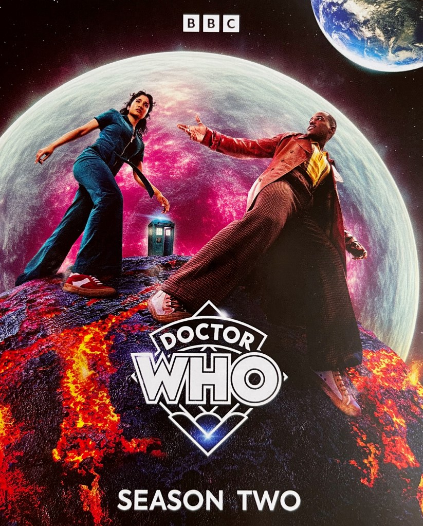 Front side of the leaflet inside the Doctor Who Season 2 Blu-ray set. It depicts Ncuti Gatwa's Doctor with companion Belinda Chandra, viewed from a low angle so they appear to be towering giants, with the Tardis very small in the background. They're standing on a meteor, covered in cracks that reveal lava beneath the surface, as it heads towards a planet or moon that has red patches on it as if it's already been heavily damaged.