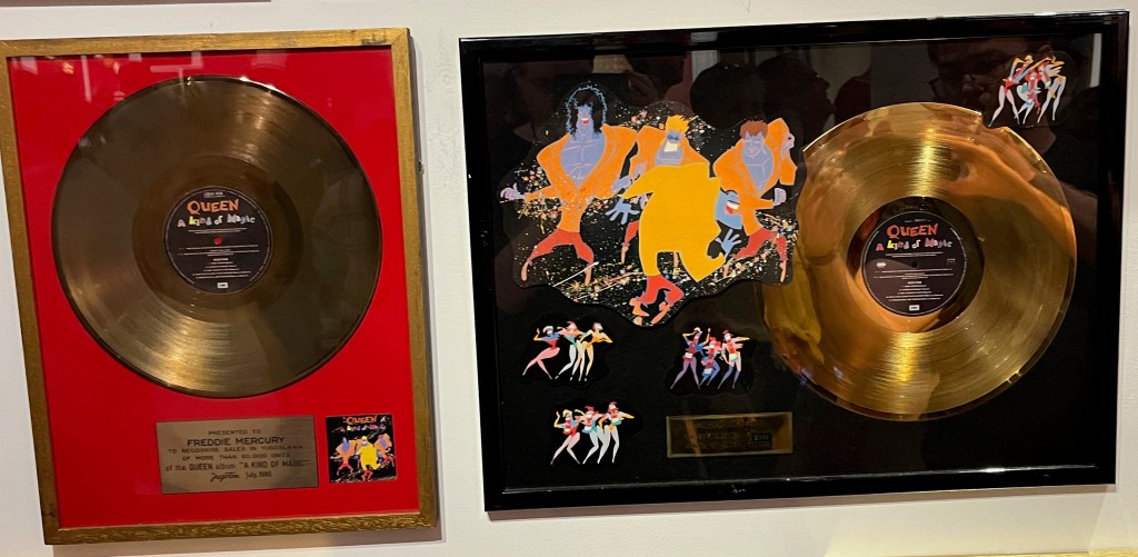 Two disc awards for the album A Kind Of Magic. Both cases contain a large gold disc, but the case on the right is wider, and to the left of the disc are the colourful caricatures of the band members from the album cover, above much smaller images in the same style showing three groups of three people dancing.