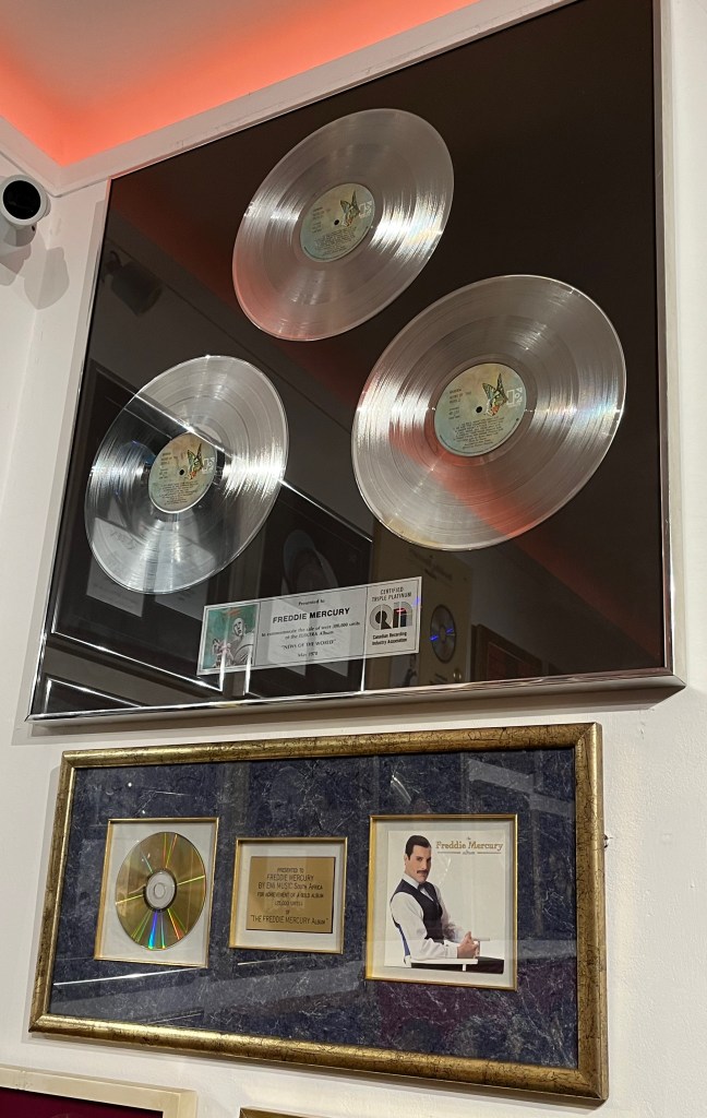 Sales award for the Queen album News Of The World, with 3 silver discs in a large case. Below that is a sales award for The Freddie Mercury album in a much smaller horizontal case, with a CD on the left and the album cover on the right, either side of the plaque in the centre.