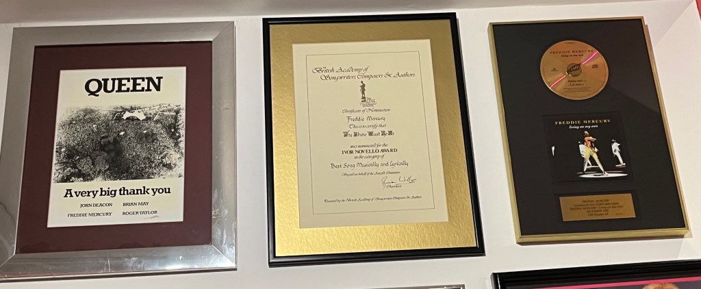 Black and white aerial photo of the crowd at Queen's Hyde Park concert in 1976, with text below that says a very big thank you, as well as the names of the four band members. To the right of the photo is an Ivor Novello certificate for The Show Must Go On and a disc award for Living On My Own.